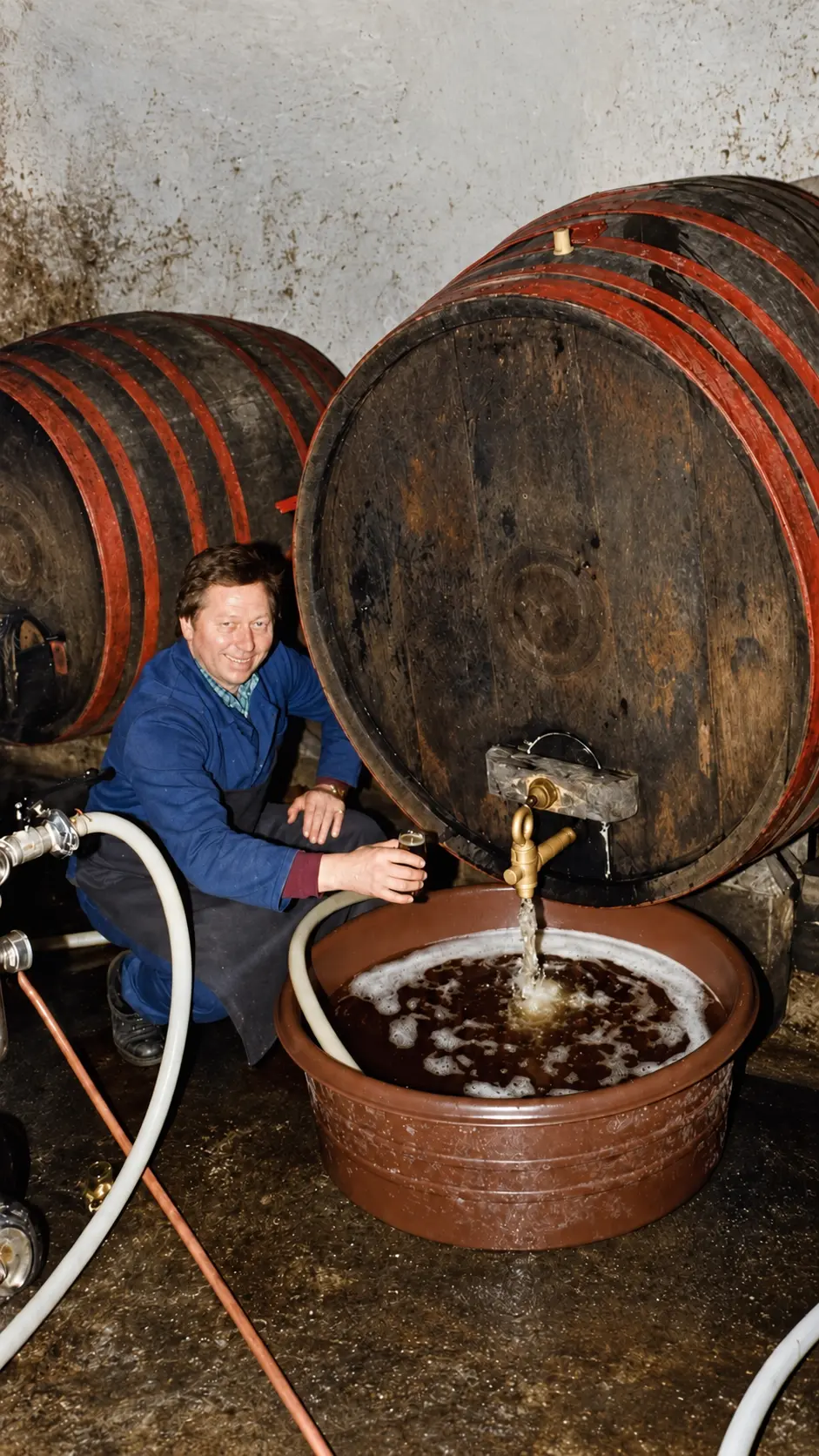 Moški v modri jopič in črnem predpasniku toči pijačo iz velikega lesenega soda v kleti
